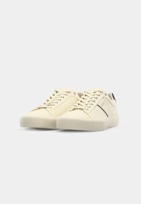 Light beige sneakers with a textured leather upper, flat lace-up design, round toe, rubber sole, and black accent near the heel.