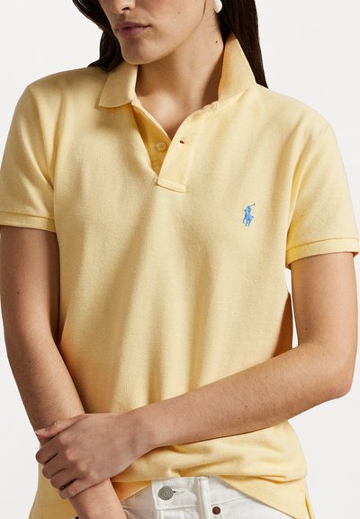 Polo shirt in light yellow, made from textured fabric, with a three-button placket and a blue embroidered logo on the chest.