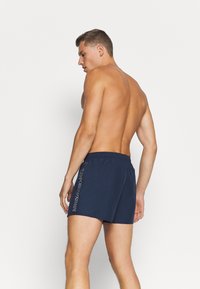 Bañador azul marino con cinturilla elástica, que presenta un estampado de logo en el lateral y una textura suave. Diseñado para la comodidad y la facilidad de movimiento.