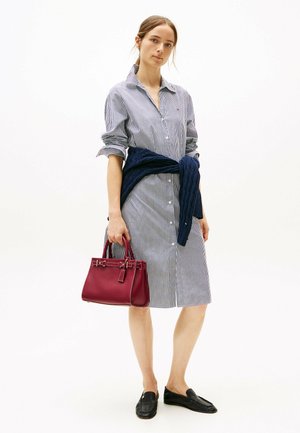 Vestito camicia a righe, con bottoni, di colore blu chiaro e bianco, abbinato a un maglione lavorato a maglia navy legato in vita. Tiene in mano una borsa bordeaux.