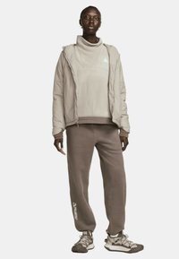 Giacca isolata beige con zip frontale, indossata sopra un top in pile grigio chiaro e pantaloni della tuta coordinati. Tessuto strutturato con dettagli del logo.