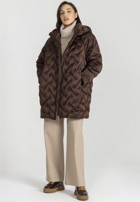 Brązowa pikowana kurtka puchowa z kapturem, z zamkiem błyskawicznym i bocznymi kieszeniami. Nosi ją na beżowym golfie i dopasowanych spodniach.