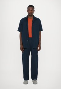 Marineblaue kurzärmlige Button-Up-Bluse über einem orangefarbenen Rundhalsausschnitt-T-Shirt, kombiniert mit marineblauen Hosen und grauen Sneakers. Glatter Stoff und taillierte Passform.