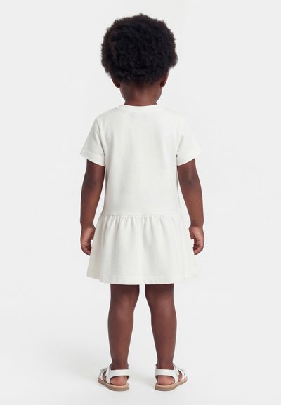 Παιδί με κατσαρά μαλλιά, ντυμένο με λευκό φόρεμα κοντών μανικιών και λευκά σανδάλια, στέκεται γυρισμένο με την πλάτη σε μια απλή φόντο.