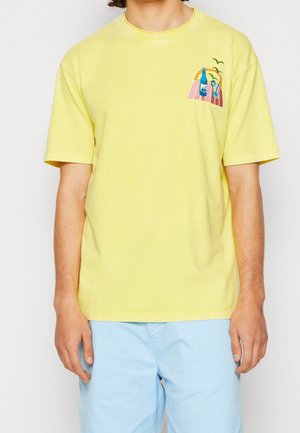 Camiseta amarilla de manga corta con escote redondo. Presenta un diseño bordado colorido en el lado izquierdo del pecho, que representa botellas y palmeras.