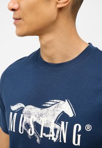 Námořnická modrá bavlněná tričko s velkým bílým potiskem koně a slovem "MUSTANG" psaným tučnými písmeny. Klasický kulatý výstřih.