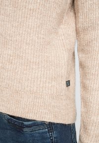 Beige pletený svetr s žebrovanou texturou. Výrazný boční štítek. V kombinaci s tmavě modrými džíny, které ukazují tradiční prošívání a klasický střih.