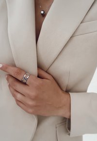 Blazer beige su misura con una finitura testurizzata. Anello d'argento con un design geometrico sul dito, abbinato a una delicata collana d'argento.