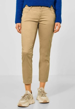 Pantalons beige fuselés avec une texture lisse, comportant cinq poches et une fermeture à bouton, associés à des baskets de couleur neutre.