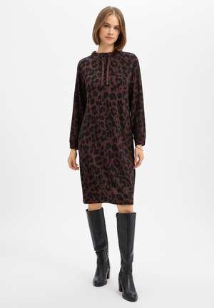 Donna con capelli corti che indossa un vestito marrone e nero a stampa leopardata a maniche lunghe e stivali neri con tacco alti fino al ginocchio, in piedi davanti a uno sfondo bianco.
