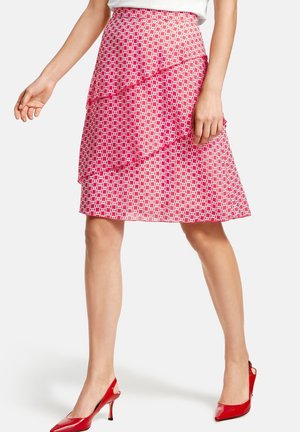 Vrouw draagt een knielange roze rok met een wit geometrisch patroon en gelaagde ruches, gecombineerd met rode hoge hakken.