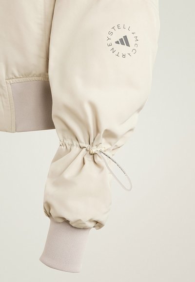 Beige bomberjack met elastische manchetten en trekkoord, voorzien van een cirkelvormig grijs logo op de mouw, gladde stof en geribbelde textuur.