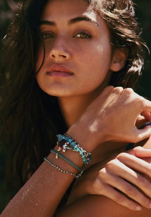 Des bracelets colorés en perles et en tissu sont portés sur un bras ayant un teint de peau bronzé texturé et de la sable visible.