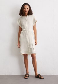 Vestito camicia in lino beige chiaro con maniche corte, chiusura frontale a bottoni e cintura in vita, caratterizzato da un orlo curvo e spacchi laterali. Sandali marroni.