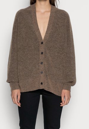 Femme portant un cardigan marron ample boutonné à manches longues et un pantalon noir sur un fond uni.