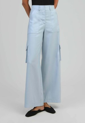 Femme portant un pantalon cargo large bleu clair avec de grandes poches avant et latérales, associé à des chaussures plates noires à enfiler.