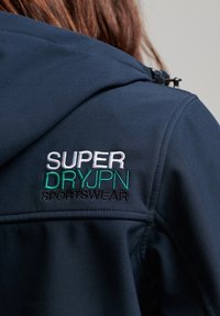 Mörkblå sportjacka i mjukt tyg med en broderad logotyp i vitt och grönt: "SUPER DRYJPN SPORTSWEAR" på baksidan.