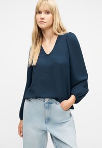 Νεαρή γυναίκα με μακριά ξανθά μαλλιά, που φοράει μπλε μπλούζα με V λαιμόκοψη και ανοιχτό μπλε τζιν, με το ένα χέρι στην τσέπη, μπροστά σε λευκό φόντο.