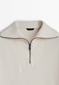 Maglione zip-up color crema con colletto ripiegato e zip nera parzialmente aperta sul colletto.