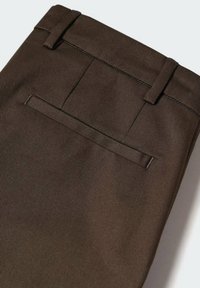 Detailní záběr na tmavě hnědé kalhoty se štíhlým pasem a jednou zadní kapsou s jemným švem.