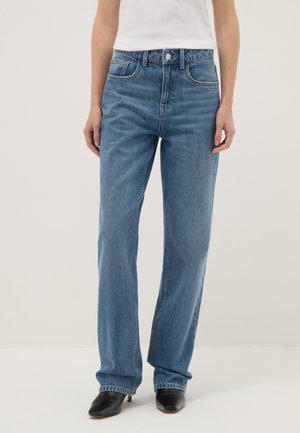 Persona que lleva puestos vaqueros azules de pierna recta, tacones negros de punta fina y una camiseta blanca metida por dentro, sobre un fondo claro liso.