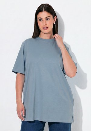 Frau mit langen dunklen Haaren, die ein lockeres hellblaues T-Shirt und dunkelblaue Hosen trägt, berührt den Kragen und steht vor weißem Hintergrund.