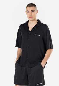 Chemise noire à manches courtes et à boutons avec un col, présentant un logo sur le côté gauche. Associée à un short en maille noir, l'ensemble est décontracté dans son design.