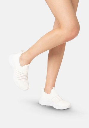 Ben iført hvide slip-on strikkede sneakers med struktureret sål, den ene fod løftet, mod en enkel lys baggrund.