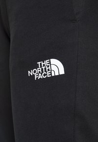 Černé tepláky vyrobené z bavlněné směsové tkaniny s bílým logem "THE NORTH FACE" na levém stehně. Hladká textura, tapered střih.