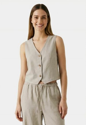 Egy nő mosolyog, világos bézs színű, gombos, ujjatlan mellényt és hozzáillő, zsinóros nadrágot visel egy egyszínű háttér előtt.