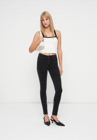 Top corto blanco con tirantes negros, combinado con jeans ajustados negros y tacones de punta negros. La modelo sostiene un pequeño bolso beige.