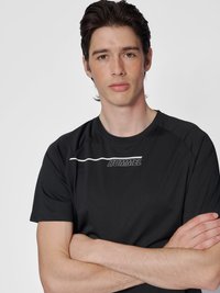 Ung mann med mørkt hår iført svart Hummel sportskjorte, med armene i kors, nøytral uttrykk, ensfarget lys grå bakgrunn.