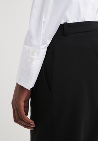 Camisa blanca de manga larga con puños abotonados, combinada con pantalones negros. La tela parece suave con un ligero brillo, mostrando un ajuste a medida.