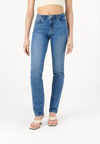 Femme portant un jean slim bleu clair et des sandales à talons ouvertes beige, debout sur un fond blanc.