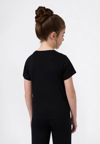 Νεαρή κοπέλα με καστανά μαλλιά συγκεντρωμένα σε μπουρνούζι, στραμμένη προς τα πίσω, φορώντας ένα απλό μαύρο κοντομάνικο μπλουζάκι και μαύρο παντελόνι, σε λευκό φόντο.