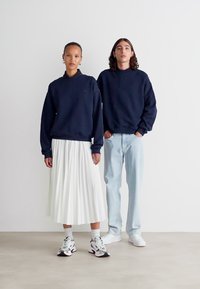 Marineblaue Sweatshirts, weißer plissierter Rock, hellblaue Jeans und gemusterte Sneakers. Minimalistisches Design mit kontrastierenden Farben und Texturen.