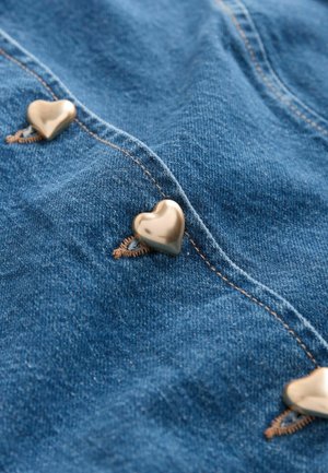 Tummansinisestä farkkukankaasta, jossa on sydämenmuotoiset kultaiset napit sauman varrella ja kontrastisia ruskeita ompeluja.