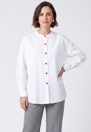 Femme portant une chemise blanche à manches longues avec des boutons rouges et un pantalon gris, debout avec une main sur la hanche devant un fond uni.