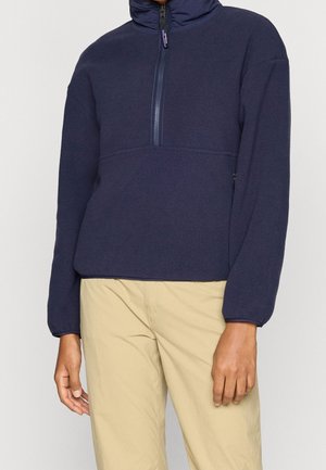 Personne portant un pull polaire bleu marine à demi-fermeture éclair et un pantalon beige clair, debout les bras détendus devant un fond uni.