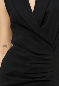 Robe noire sans manches avec col à revers et détail de tissu froncé à la taille, laissant voir une partie du bras avec un petit tatouage.