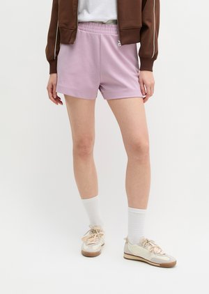 Osoba oblečená v svetlofialových elastických krátkych nohaviciach, bielych ponožkách, béžových teniskách, hnedej bunde, stojaca pred obyčajným bielym pozadím.