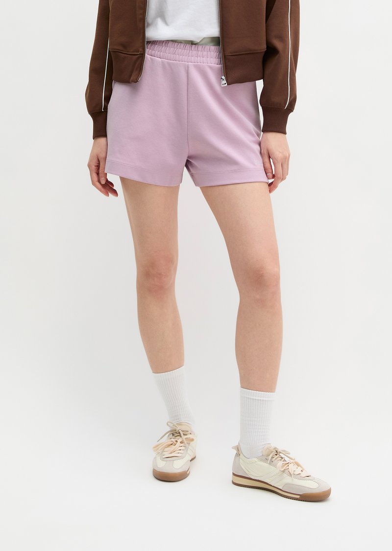 Άτομο που φοράει ανοιχτό μωβ ελαστικά σορτς με λάστιχο στη μέση, λευκές κάλτσες, μπεζ αθλητικά παπούτσια, καφέ μπουφάν, στέκεται σε ένα απλό λευκό φόντο.