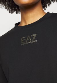 Osoba oblečená v černém crewneck tričku se zlatým logo "EA7 Emporio Armani" umístěným uprostřed pod výstřihem.