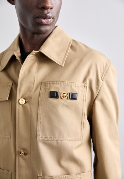 Beige canvas jacket featuring two front pockets, gold button accents, and a decorative emblem with black and gold hardware.