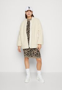 Crème nepbontjack met een ritssluiting, gedragen over een fitted jurk met luipaardprint. Model draagt witte chunky enkellaarsjes en een witte pet.