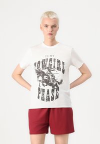 Witte katoenen T-shirt met een zwart grafisch ontwerp van een cowboy op een paard met de tekst "IN MY COWGIRL PHASE". Korte mouwen, ronde halsontwerp.