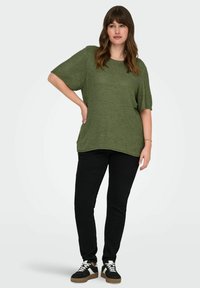 Žalias mezgamas viršus su trumpomis rankovėmis ir laisvu siluetu, derinamas su juodomis siaurėjančiomis kelnėmis ir juodais sportbačiais, turinčiais baltus ir rudus akcentus.