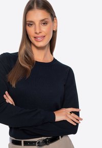 Granatowy sweter z dzianiny z długimi rękawami, okrągłym dekoldem i prążkowaną fakturą. Noszony z beżowymi spodniami i czarnym paskiem, z złotym pierścieniem na palcach.