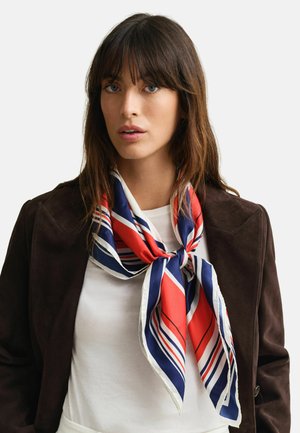 Femme aux cheveux bruns portant une veste marron foncé et une chemise blanche, accessoirisée d'une écharpe rayée rouge, blanche et bleue nouée autour du cou.