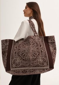 Duża brązowa torba tote z tkaniny, zdobiona misternymi białymi wzorami paisley oraz jednolitymi brązowymi akcentami, z szerokimi uchwytami ułatwiającymi noszenie.